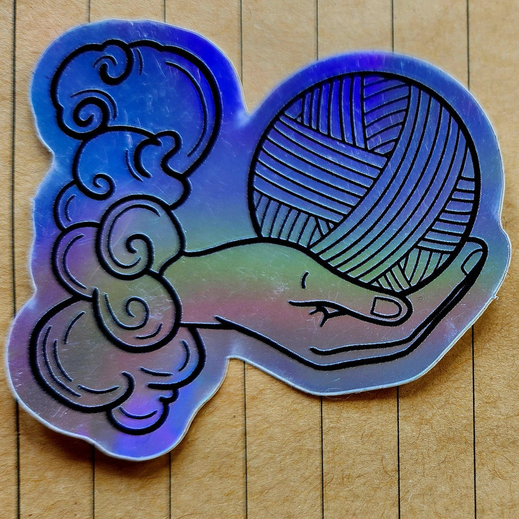 A holographic sticker featuring a hand holding a ball of yarn with a tarot card design on a wooden background.