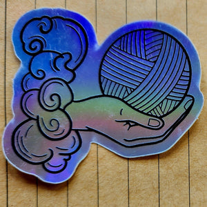 A holographic sticker featuring a hand holding a ball of yarn with a tarot card design on a wooden background.