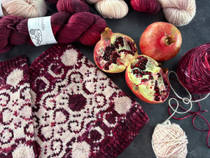 Knitted cowl with burgundy and light pink pomegranate and filigree pattern, pomegranates, and yarn on a dark surface