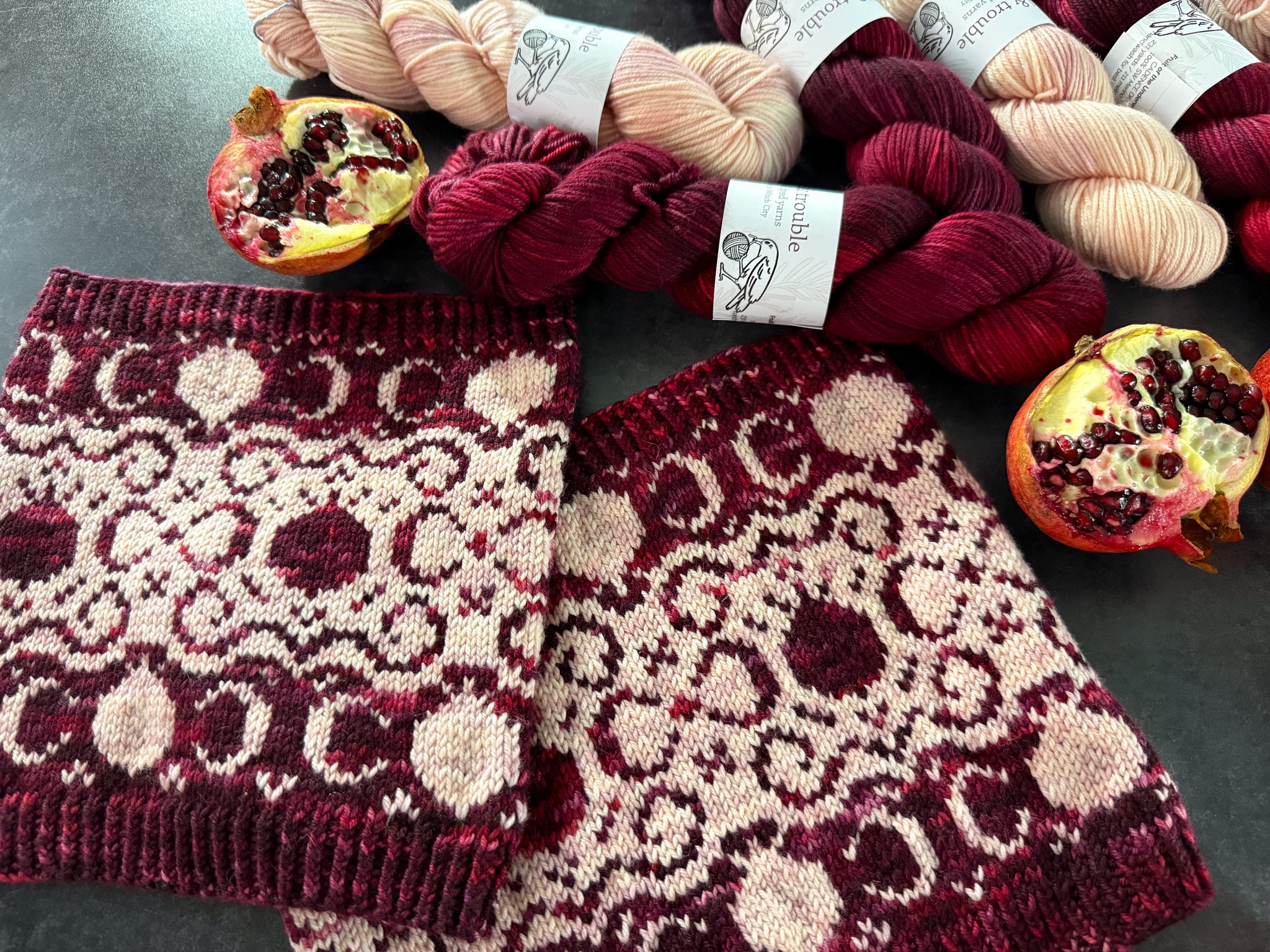 Knitted cowls with burgundy and light pink pomegranate and filigree pattern, pomegranates, and skeins of yarn on a dark surface