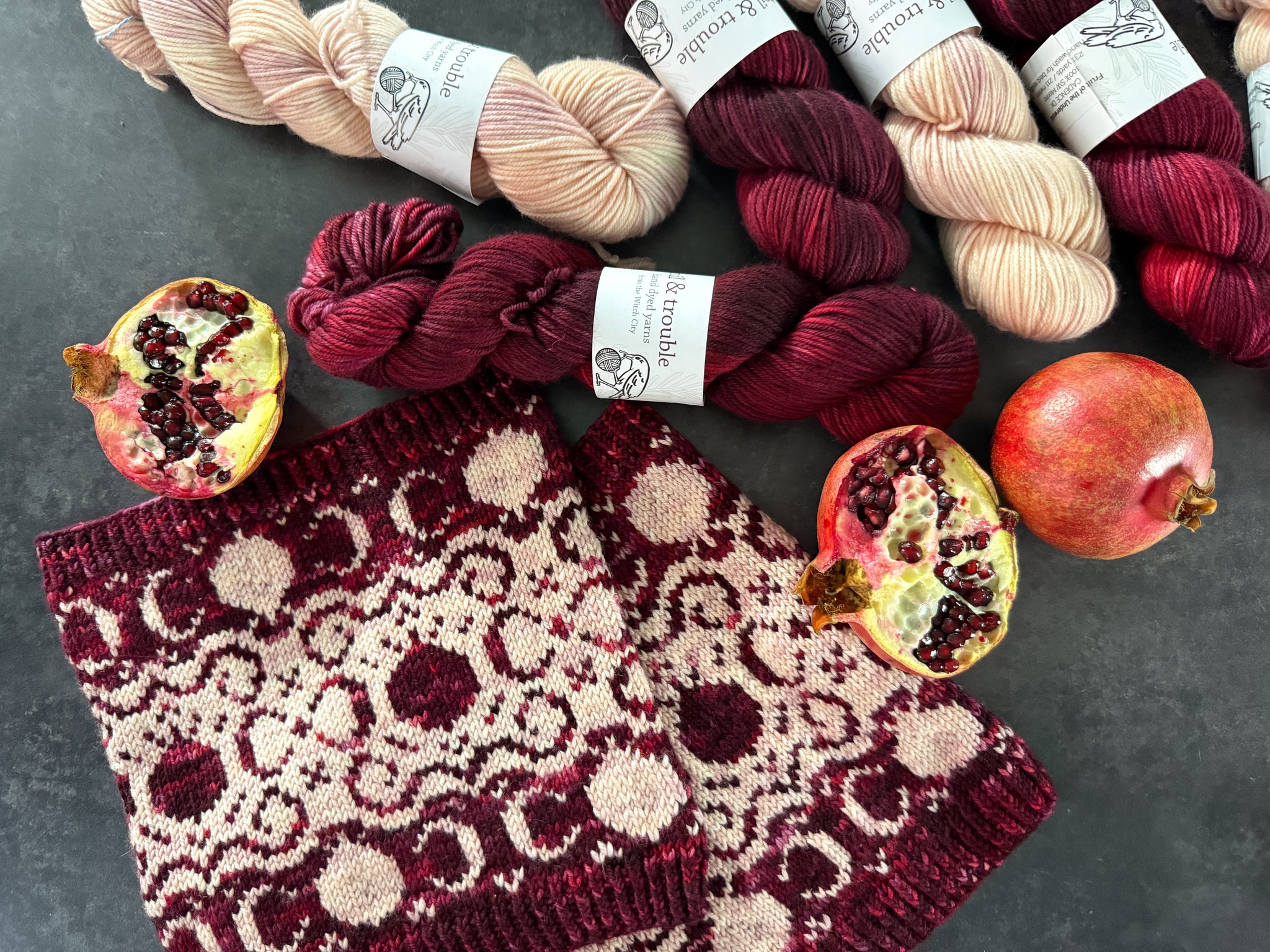 Knitted cowls with burgundy and light pink pomegranate and filigree pattern, pomegranates, and yarn on a dark surface