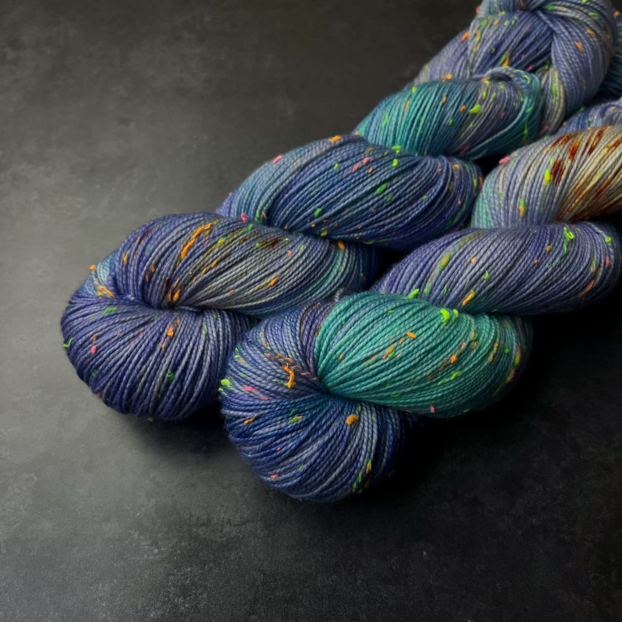 Skeins of purple and aqua tweed sock yarn with orange speckles and colorful neon slubs, displayed on a dark textured surface.