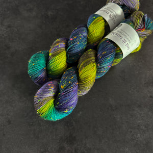Skeins of aqua, cool purple, and bright green tweed sock yarn with colorful neon slubs, displayed on a dark textured surface.