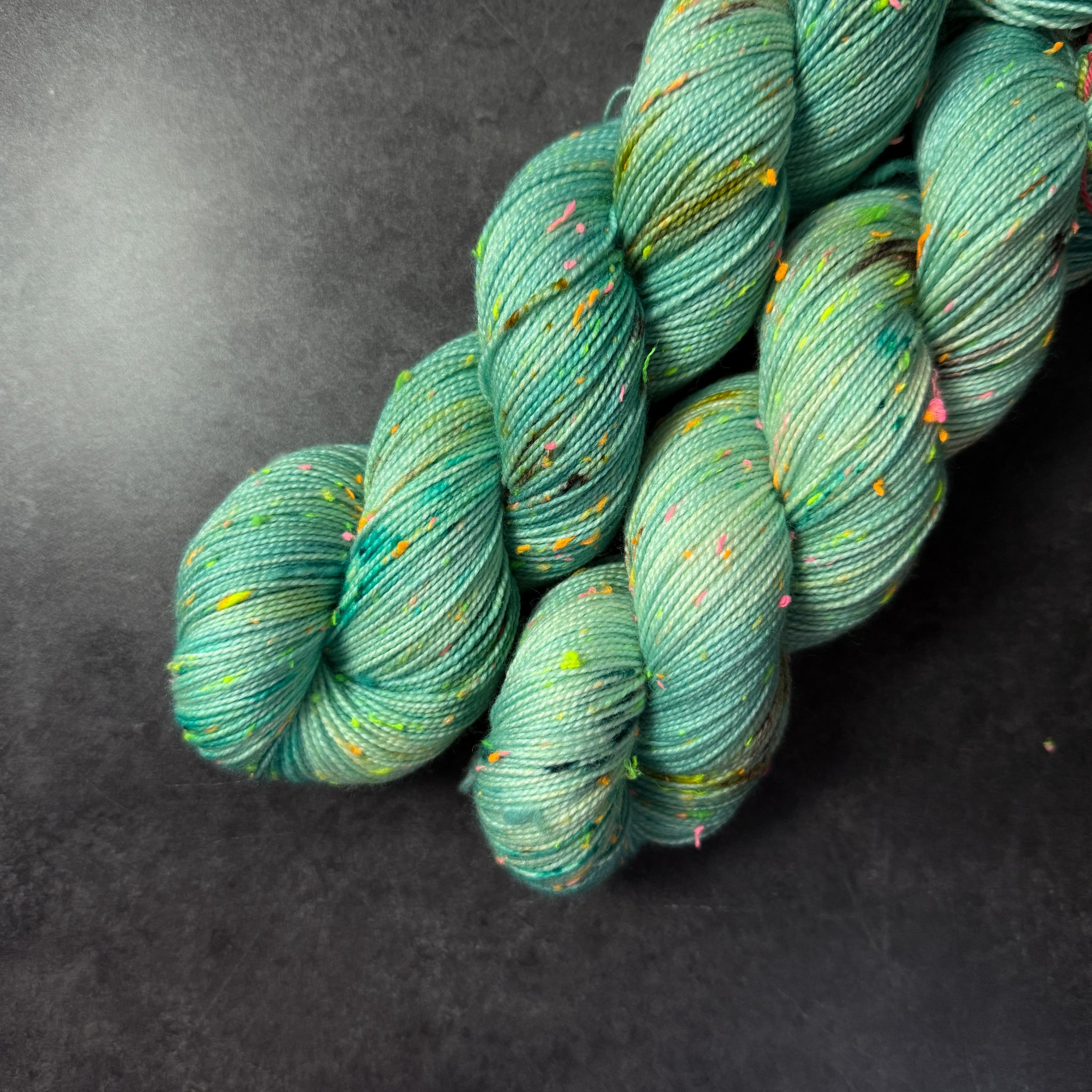 Skeins of turquoise tweed sock yarn with colorful neon slubs, displayed on a dark textured surface.