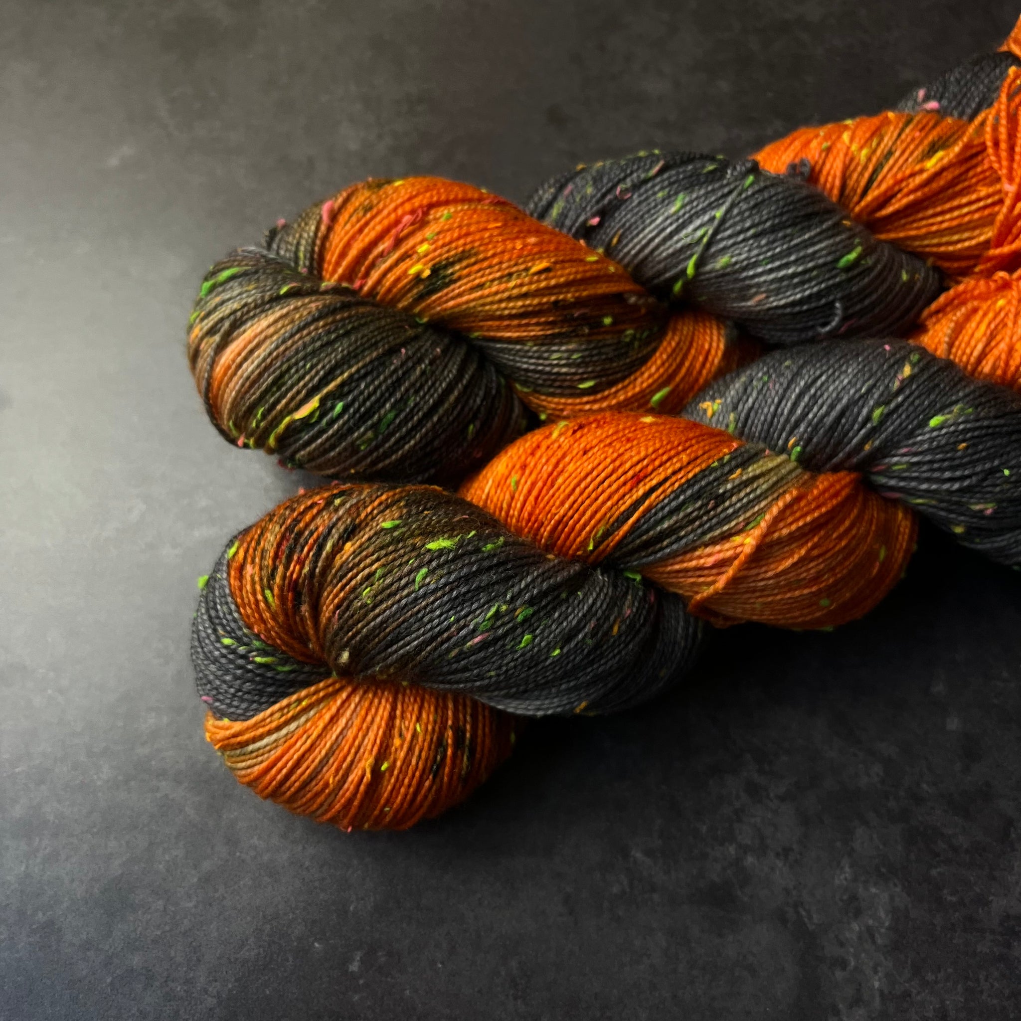 Skeins of black and orange tweed sock yarn with colorful neon slubs, displayed on a dark textured surface.