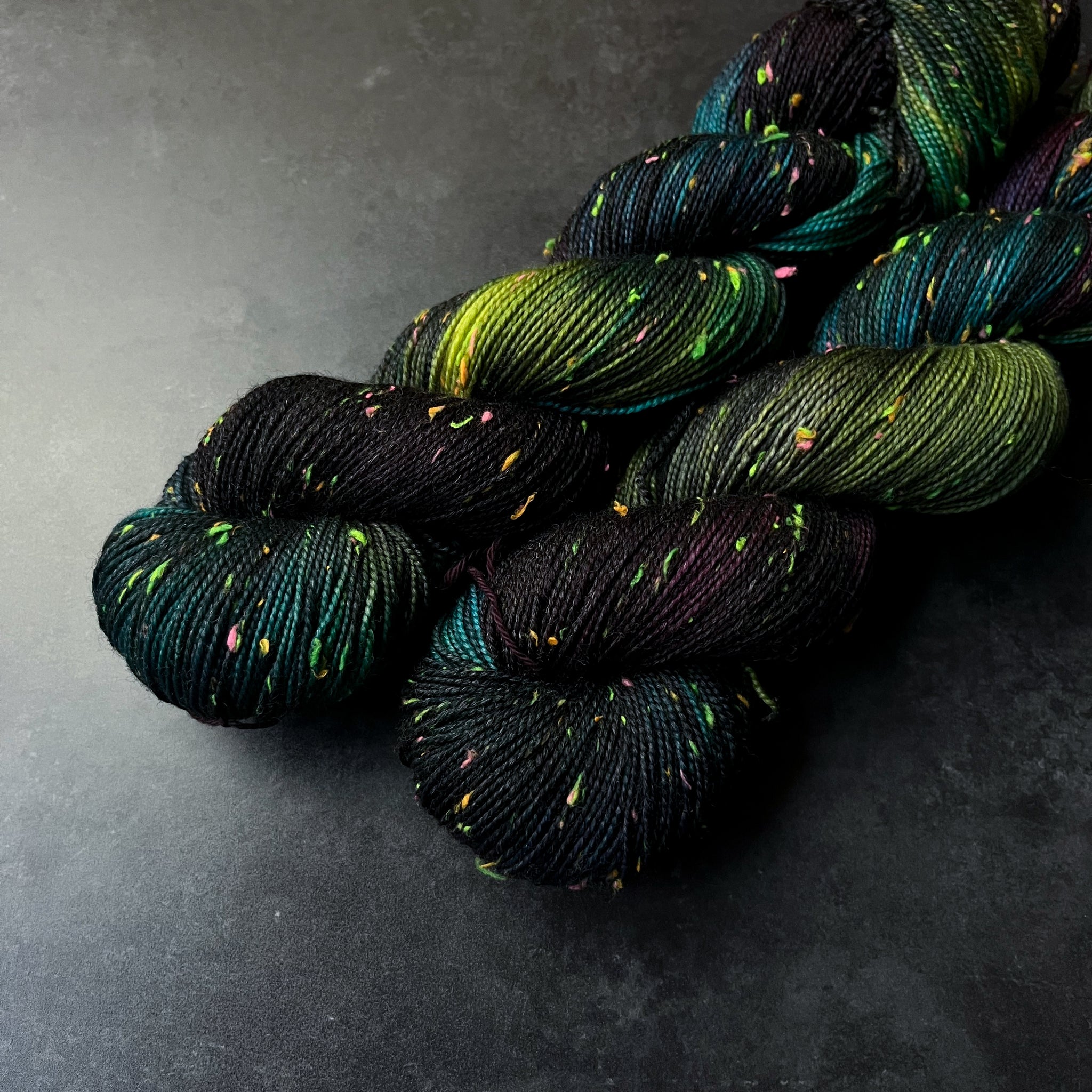 Skeins of black, dark turquoise, purple, and acid green tweed sock yarn with colorful neon slubs, displayed on a dark textured surface.