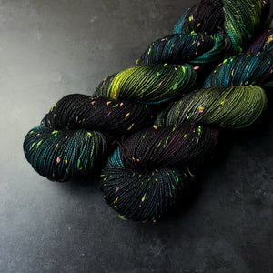 Skeins of black, dark turquoise, purple, and acid green tweed sock yarn with colorful neon slubs, displayed on a dark textured surface.