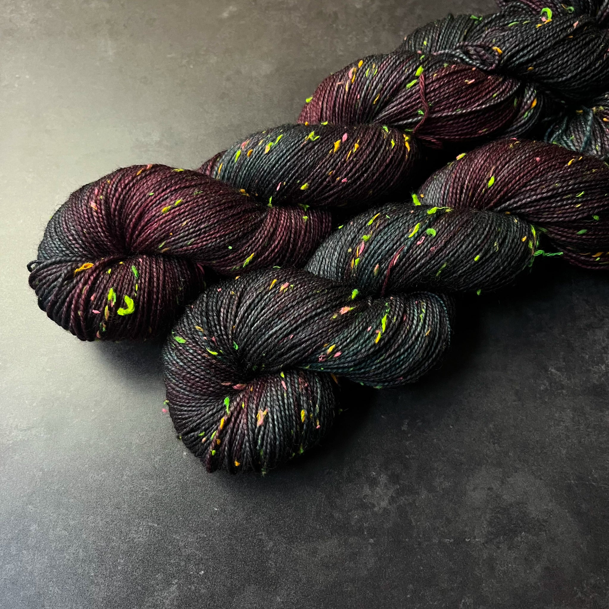 Skeins of very dark teal and burgundy tweed sock yarn with colorful neon slubs, displayed on a dark textured surface.