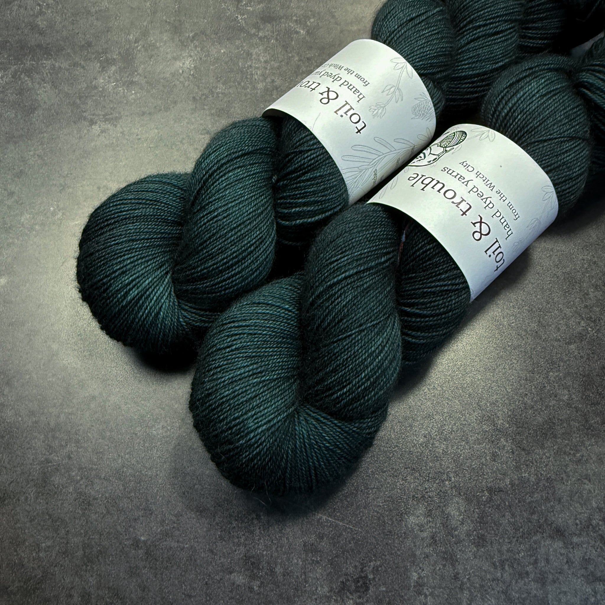 Two skeins of dark teal yarn with labels on a gray surface