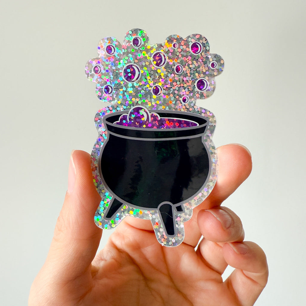 Hand holding a colorful, glittery black cauldron stickert against a white background