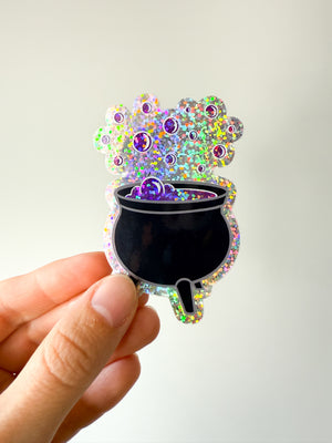 Hand holding a colorful sticker of a cauldron with bubbles against a white background